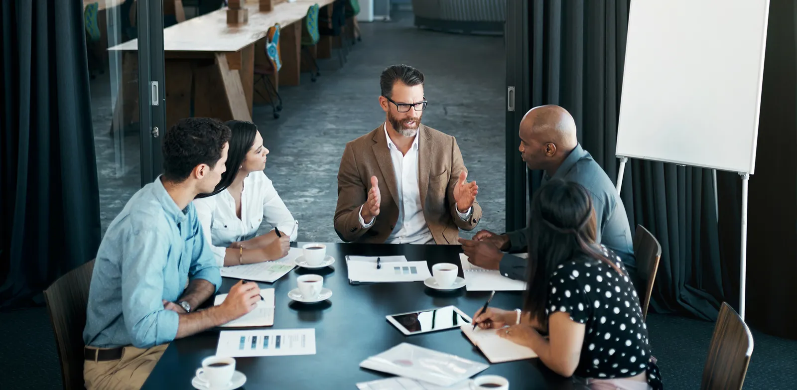 Cinq personnes sont en réunion autour d'une grande table où sont posés des tasses de café et des documents de travail.